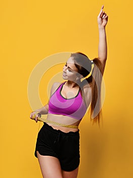 Beautiful sports woman measure her waist with hand pointing finger up on yellow background
