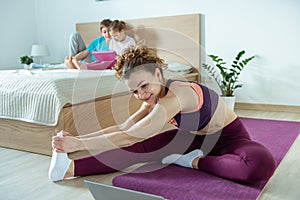 Beautiful sporty woman making fitness or yoga training online using laptop in bedroom. Children playing with tablet on bed on