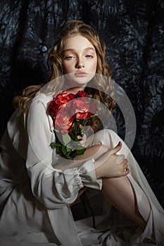 Beautiful spring girl is sitting on the floor with rose flowers in her hands. Woman in a white dress is dreaming, a romantic image