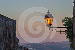 A beautiful street lamp lights up against the background of a remote hilly residential development at dusk. Symbol of the onset of