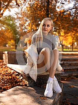 Beautiful student girl with perfect legs sitting on the parapet in the autumn park in white sneakers and coat