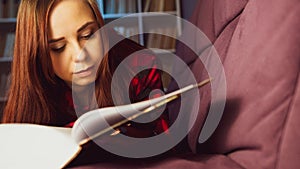 A beautiful student reading a big book. A thoughtful young woman lying on the sofa with a book and reading in a dark