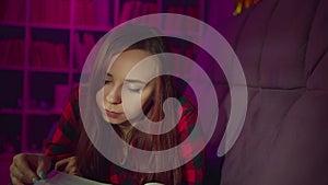 A beautiful student reading a big book. A thoughtful young woman lying on the sofa with a book and reading in a dark