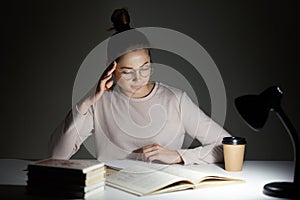 Beautiful student sits at white desk against dark background, tries to concentrate under light of lamp, wears casual jamper and