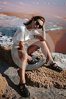 Beautiful, stunning and stylish young woman is sitting and posing on the rocks. Countryside landscape, nature, water and stones