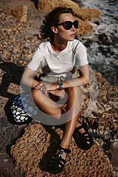 Beautiful, stunning and stylish young woman is sitting and posing on the rocks. Countryside landscape, nature, water and stones