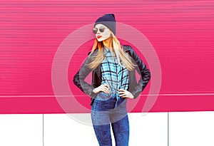 Beautiful stylish blonde woman in rock black style jacket, hat posing on city street over colorful pink wall