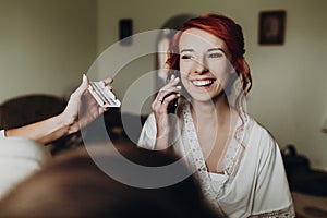 Beautiful stylish bride with red hair talking on phone and smiling, wearing robe,  getting make up done by professional, morning