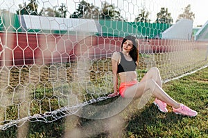 Beautiful stylish brunette woman in black top and pink shorts sitting on the lawn near a football goal at the stadium at sunset.