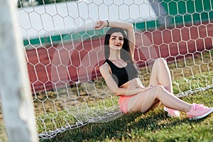 Beautiful stylish brunette woman in black top and pink shorts sitting on the lawn near a football goal at the stadium at sunset.