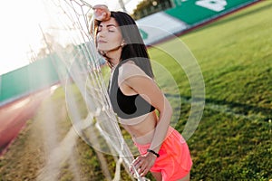 Beautiful stylish brunette woman with closed eyes in black top and pink shorts stands near a football goal at the stadium at