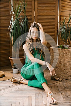 A beautiful stylish Caucasian girl with long hair and professional makeup in a green jumpsuit sits near tropical plants