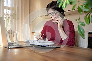Beautiful stylish elderly woman talking on a mobile phone while working at home with a laptop