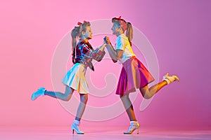 Beautiful, stylish girls, children in retro clothes dancing lindy hop against pink studio background in neon light