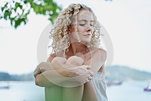 Beautiful stylish glamorous young blonde woman with curly hair sitting on the beach with bare shoulders on the beach