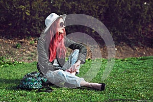 Beautiful stylish red haired fashion hipster model woman sitting outdoors on green grass at park wearing sunglasses, hat and black