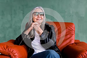 Beautiful stylish senior woman with long straight gray hair, wearing eyeglasses and black leather jacket, sitting in