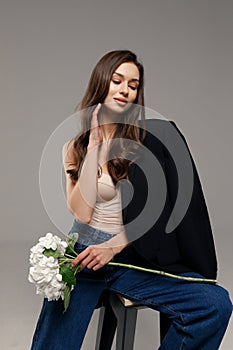 A beautiful stylish smiling girl in blue jeans and a black jacket sits on a chair on a gray background and holds white flowers in