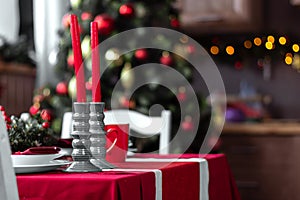 Beautiful and stylish table setting with Christmas decorations in living room, red candles and blurred background with fir tree