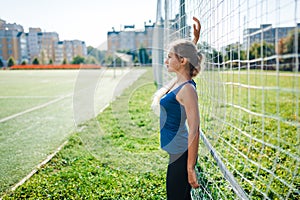 Beautiful stylish woman in a blue shirt and leggings stands near a football goal at the stadium at sunset. Beautiful sunlight. A g