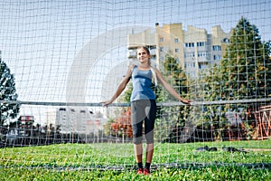 Beautiful stylish woman in a blue shirt and leggings stands near a football goal at the stadium at sunset. Beautiful sunlight. A g