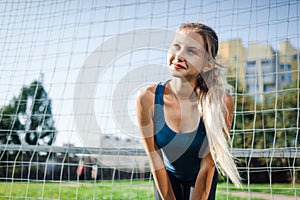 Beautiful stylish woman in a blue shirt and leggings stands near a football goal at the stadium at sunset. Beautiful sunlight. A g