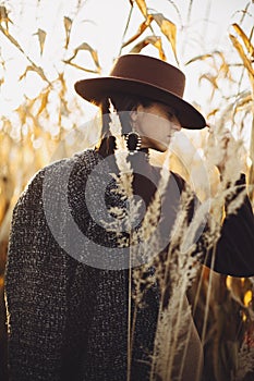 Beautiful stylish woman in brown hat and vintage coat posing in autumn maize field in sunset light. Portrait of fashionable young