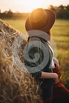 Beautiful stylish woman in hat looking at sunset light at haystacks in summer field.Tranquility