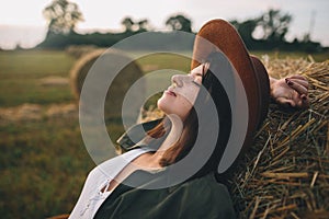 Beautiful stylish woman with herb in mouth relaxing on haystack in summer evening field. Tranquility
