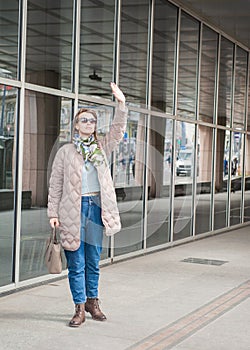 Beautiful stylish woman with sunglasses and bag walking near building with mirror wall on the street and waving her hand hello