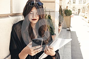 Beautiful stylish woman using phone and holding magazine in city