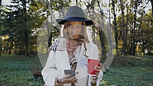 Beautiful stylish woman in the white coat and hat walking in the park with coffee and smart phone.