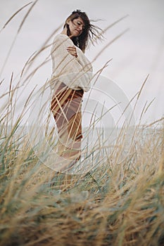 Beautiful stylish woman with windy hair in knitted sweater posing among wild grass. Carefree moment, stylish image. Fashionable