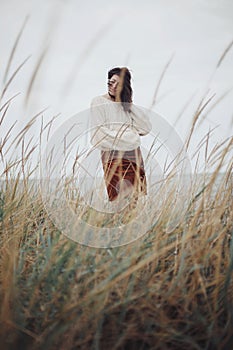 Beautiful stylish woman with windy hair in knitted sweater posing among wild grass. Carefree moment, stylish image. Fashionable