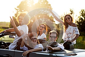 Beautiful stylish young girls and guys in sunglasses are sitting and laughing in a black cabriolet on a sunny day.