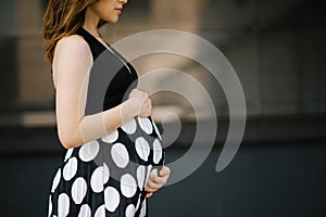 A beautiful stylish young pregnant woman in a black dress with white polka dots holds her hands on her stomach. Happy waiting for