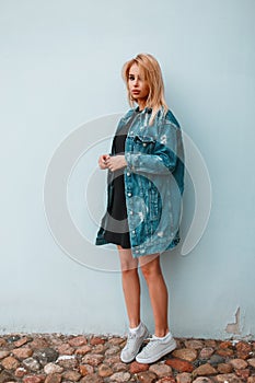 Beautiful stylish young woman blonde in a fashionable long denim jacket in a black trendy dress in sneakers near the vintage wall