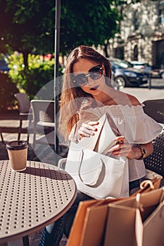 Beautiful stylish young woman with shopping bags chilling in outdoor cafe on city street and checking her purchases.