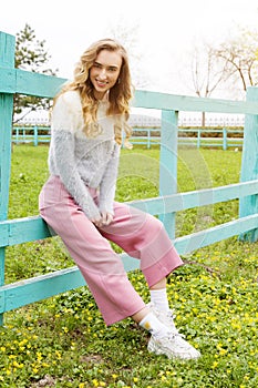 Beautiful stylish young woman sitting on a blue fence on green grass - fashion and style concept