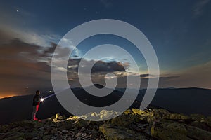 Beautiful summer night in mountains. Profile silhouette of young tourist hiker man with flashlight standing alone on rocky mountai