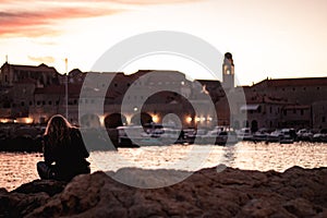 Beautiful sunset over the old town of Dubrovnik. Small local harbour in front of the city, woman in black clothes sitting on a