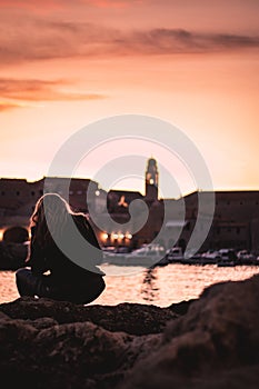 Beautiful sunset over the old town of Dubrovnik. Small local harbour in front of the city, woman in black clothes sitting on a