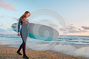 Beautiful surf girl with long hair holding surfboard go on beach at sunset or sunrise.