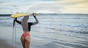 Beautiful surfer girl walking down to the beach for sunset surf session carrying blue - yellow surfboard on head with back in