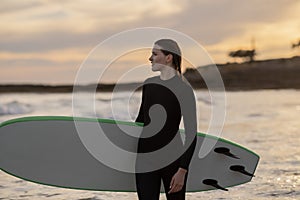 Beautiful Surfer Woman With Her Surfboard Standing On The Beach At Sunset