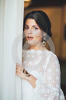 Beautiful surprised bride brunette young woman in white lace dress near window, close up portrait
