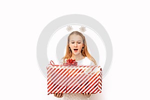 Beautiful surprised kid girl holding a Christmas red striped gift box with a ribbon and a bow in her hands. There are snowflakes