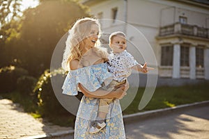 Beautiful and surprised young blonde woman in fashionable blue and yellow dress with a cute little baby boy on her hands