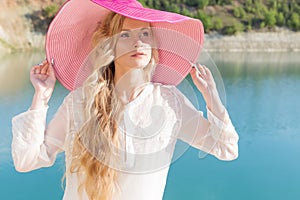 Beautiful sweet gentle girl in white dress in light big pink hat on the shore of the blue sea at sunset summer evening sun