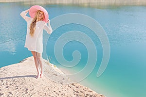 Beautiful sweet gentle girl in white dress in light big pink hat on the shore of the blue sea at sunset summer evening sun